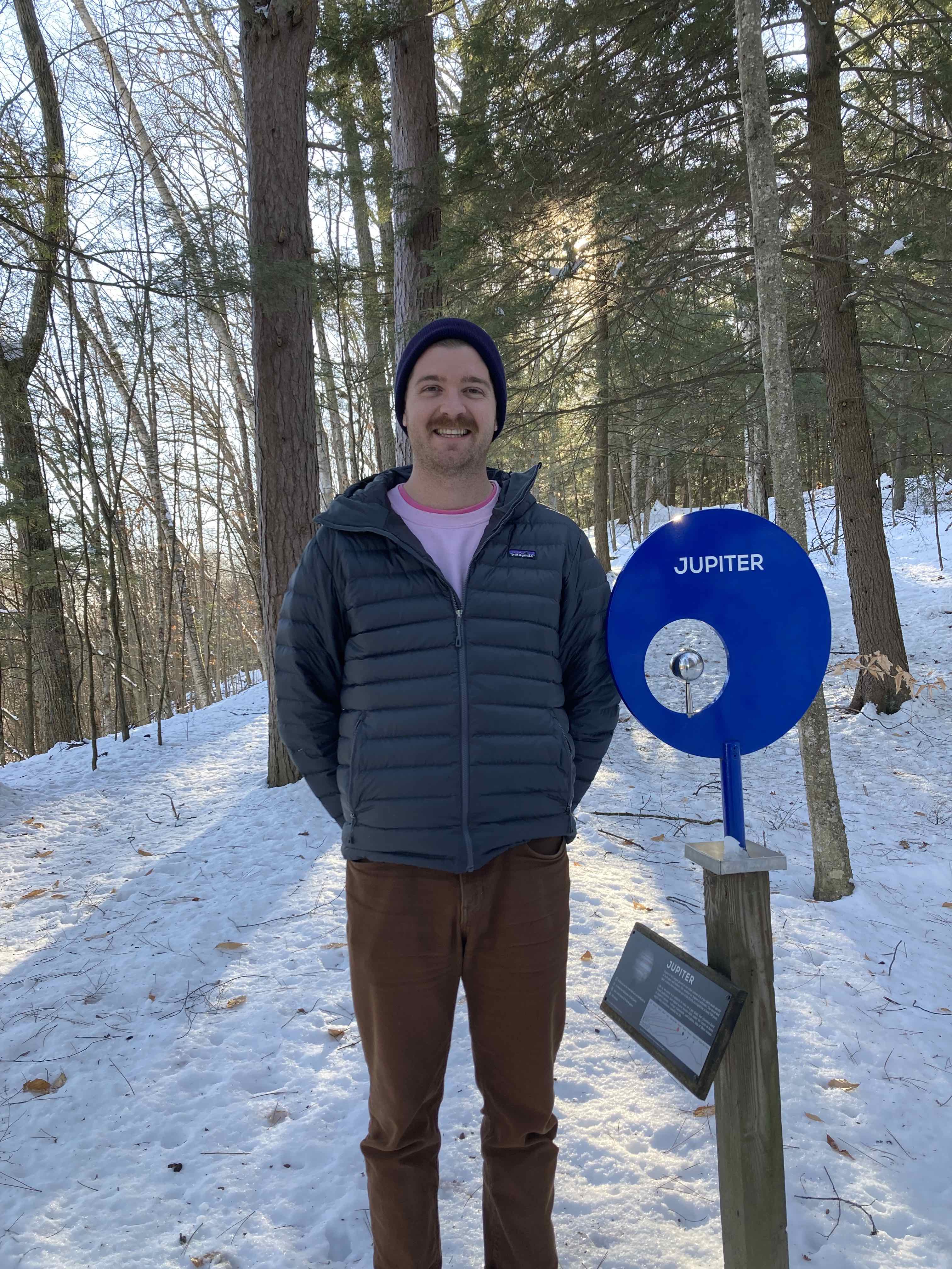 Graham standing in a snowy forest next to a post with a silvery metal ball 3 inches in diameter, labeled 'Jupiter'. Graham is white-skinned with a red mustache, wearing a blue beanie hat, a grey jacket, and brown pants.